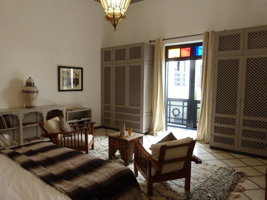 Una habitación con una cama, una silla de madera, una mesita y una estantería. Se ve una ventana con vistas a edificios.