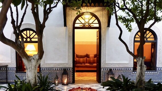 Un patio interior con suelo de baldosas, una fuente central con agua y pétalos de flores, y un acceso a una habitación con cama y lámpara.