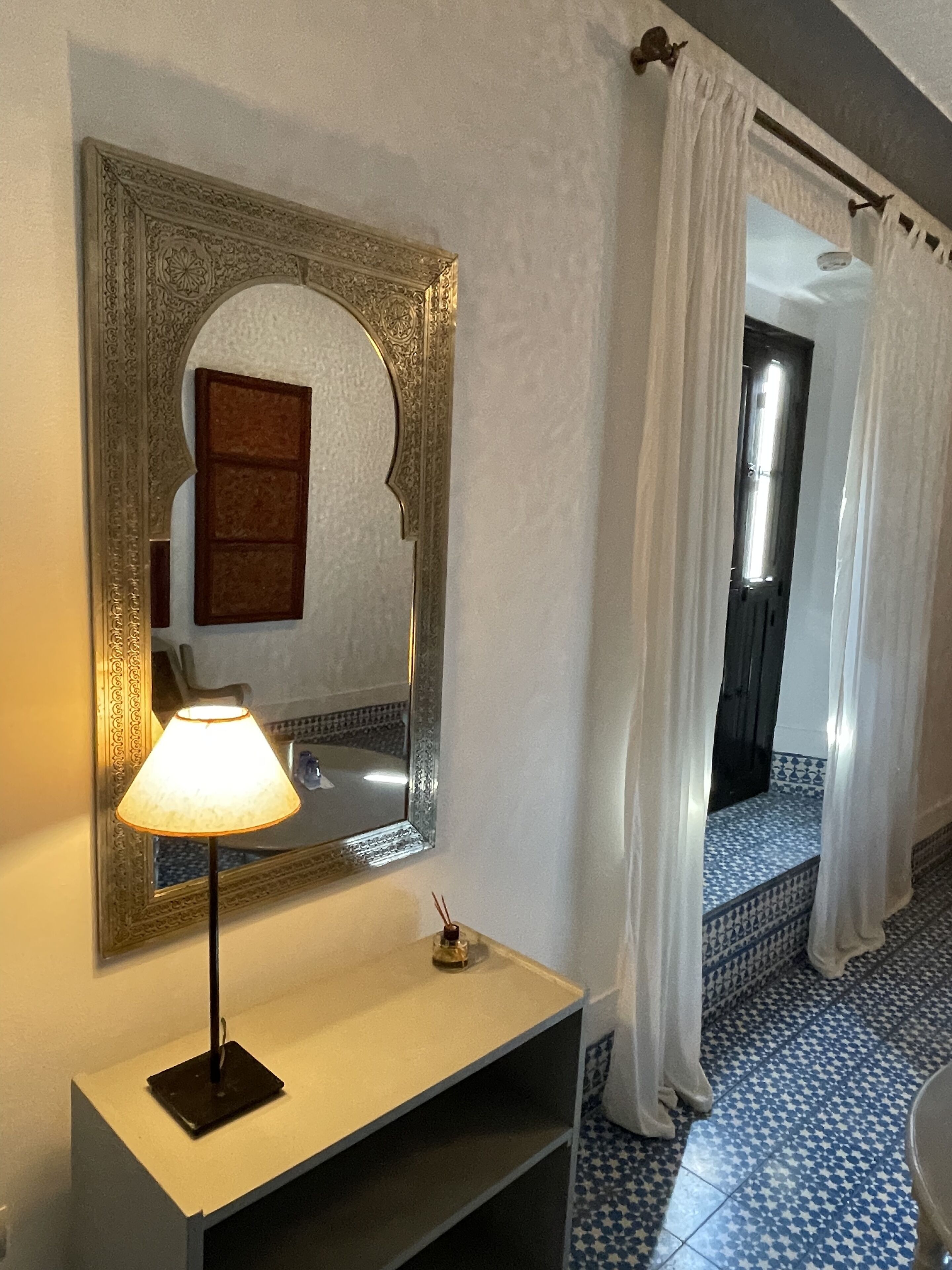 A bathroom with a mirror, a lamp, a small bottle, and a tiled floor.