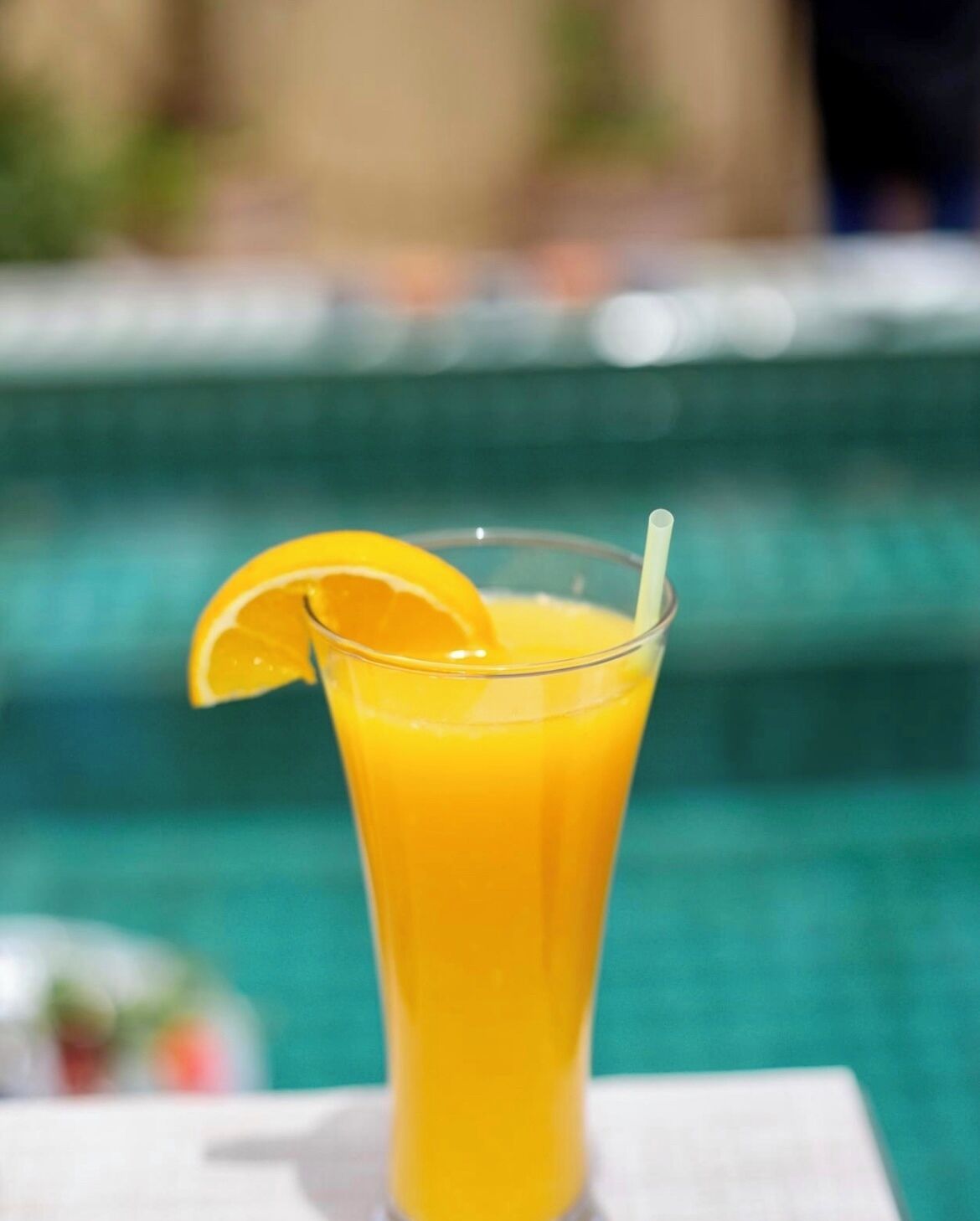 A glass of orange juice with a slice of orange and a straw, placed on a white surface near a pool.