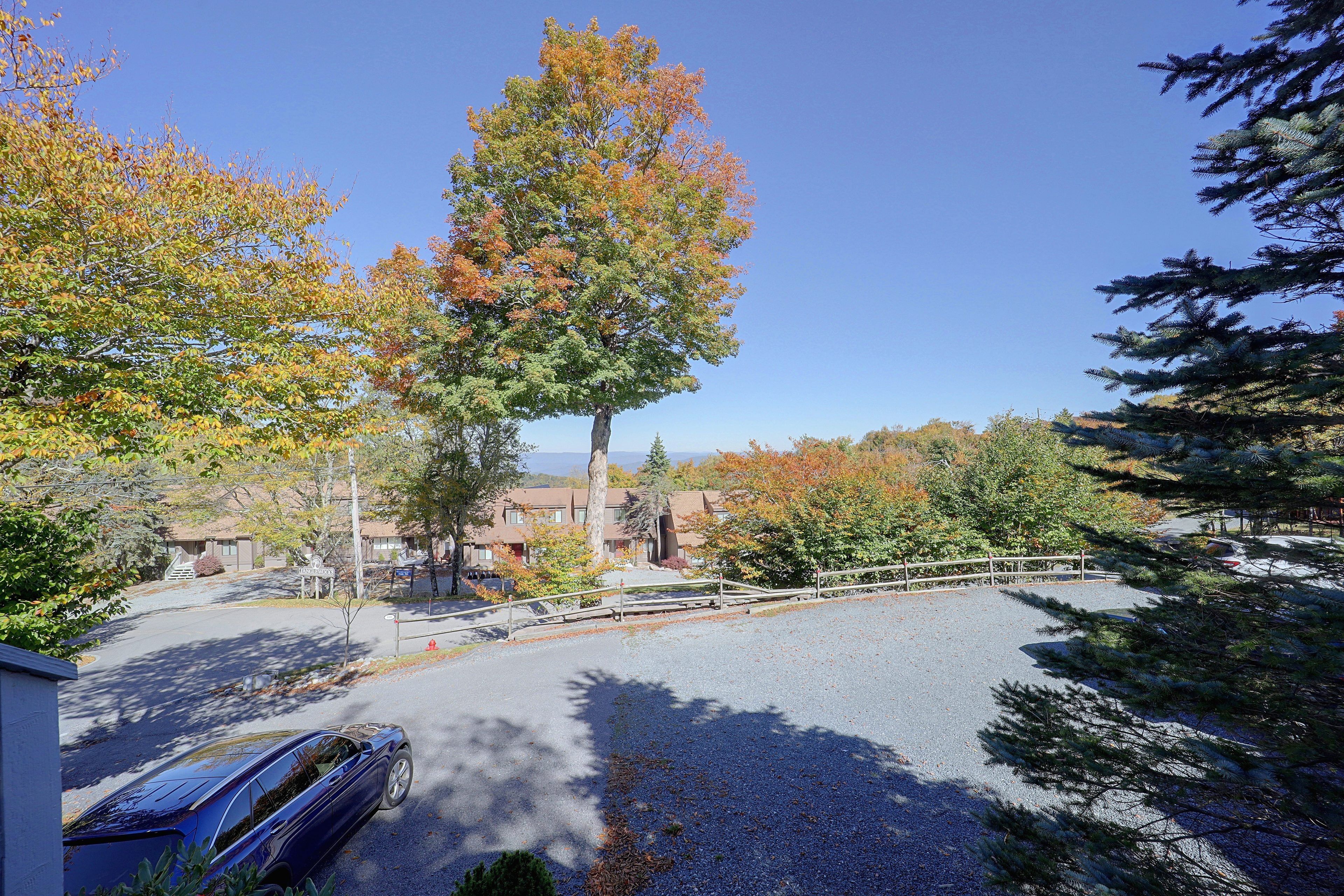 Un espace de stationnement où se trouve une voiture bleue, entourée d’arbres aux feuilles d’automne, et un bâtiment avec un toit brun en arrière-plan.