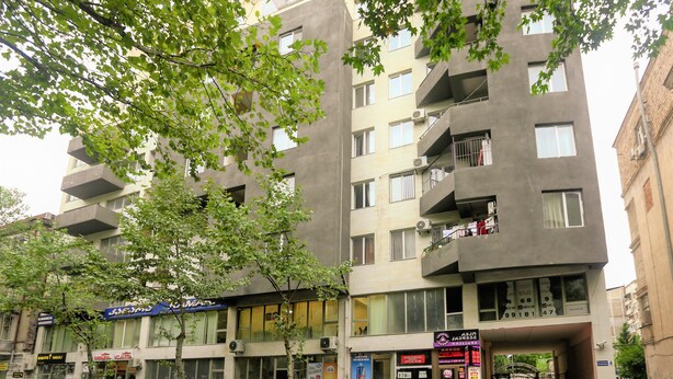 Um edifício com vários andares, varandas, lojas e carros estacionados.