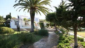 A cobblestone pathway leading to a white building with a red-tiled roof, surrounded by lush greenery and palm trees. - GARDEN APARTMENT   (Lagos)