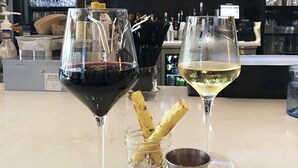 Twee wijnglazen, één met rode wijn en één met witte wijn, op een bar met een klein potje crackers en een metalen beker.