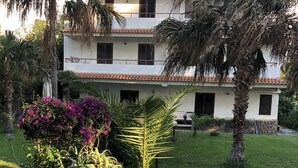 A two-story building with a tiled roof, surrounded by lush greenery including palm trees and flowering plants. - semidetached beach villa in front of eolian island near Milazzo: Tindari (furnari)