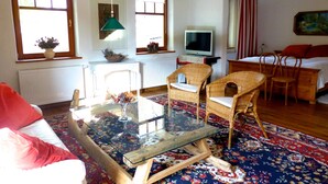 Ein gemütliches Wohnzimmer mit einem Glastisch, Korbstühlen, einem Fernseher und einem Bett mit roten Kissen.