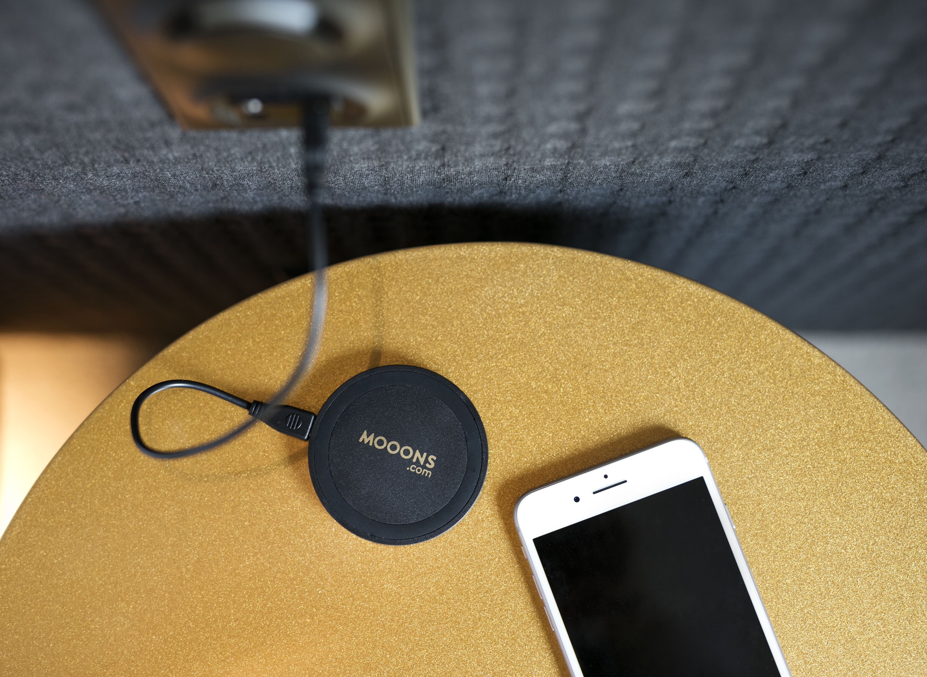 A smartphone on a golden surface with a charging cable and a black circular object labeled "MOONS.com".