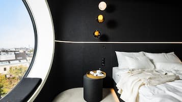 Una habitación de hotel moderna con un ventanal grande, una cama con ropa blanca, una mesita de noche pequeña con una bandeja y una lámpara fijada a la pared.
