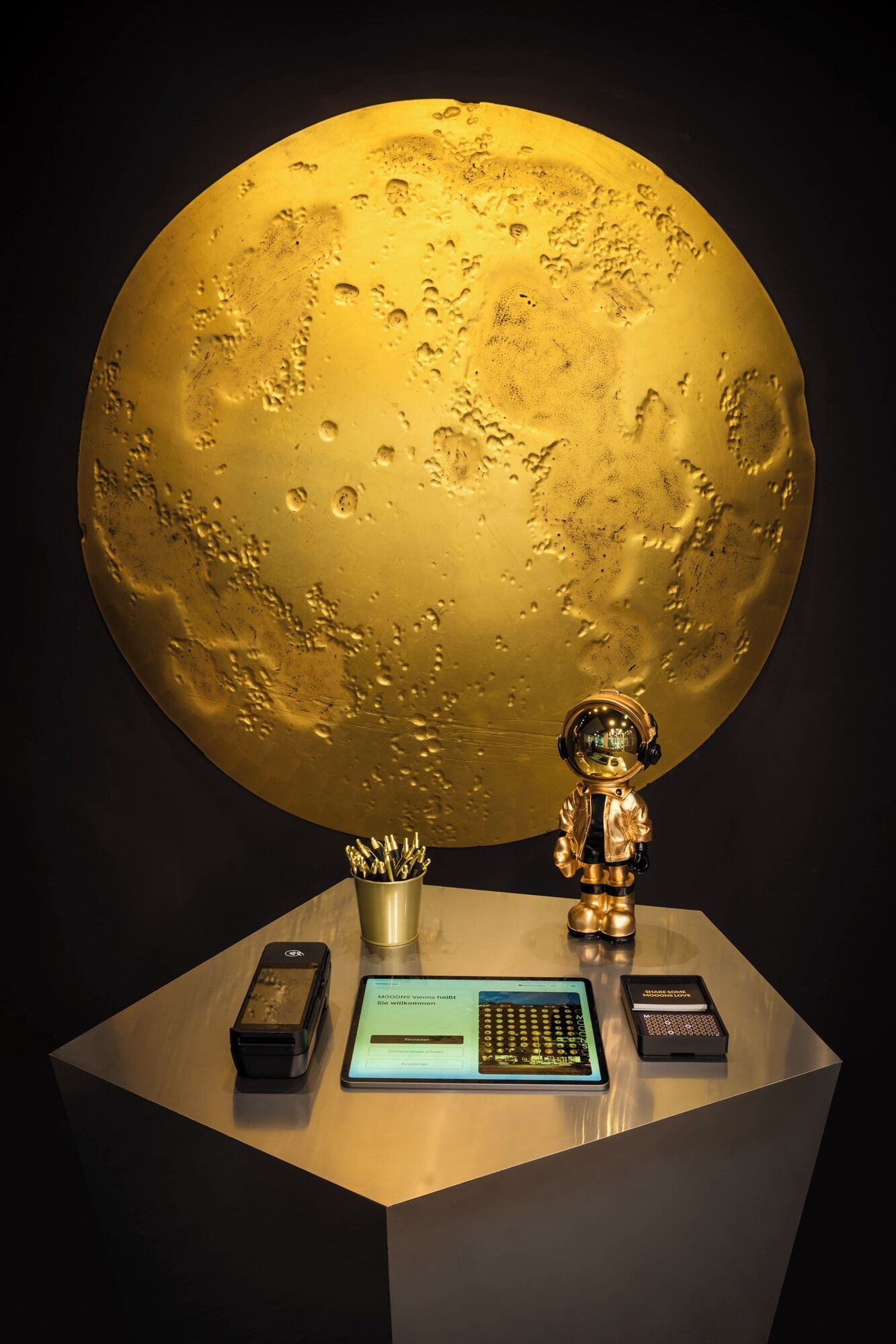 Uma mesa com uma esfera dourada grande, uma pequena figura de astronauta dourada, um tablet, uma calculadora e um recipiente de metal com canetas.