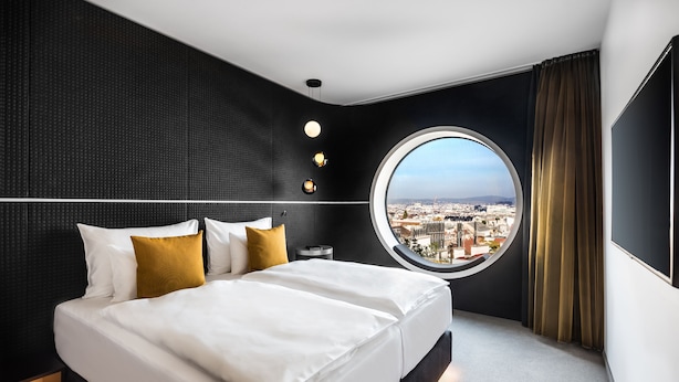 A modern hotel room with a large bed, a round window offering a city view, and a dark textured wall.