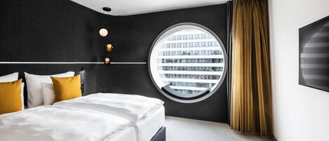Une chambre d’hôtel moderne avec un grand lit, une fenêtre ronde donnant sur la ville et un mur à la texture sombre.