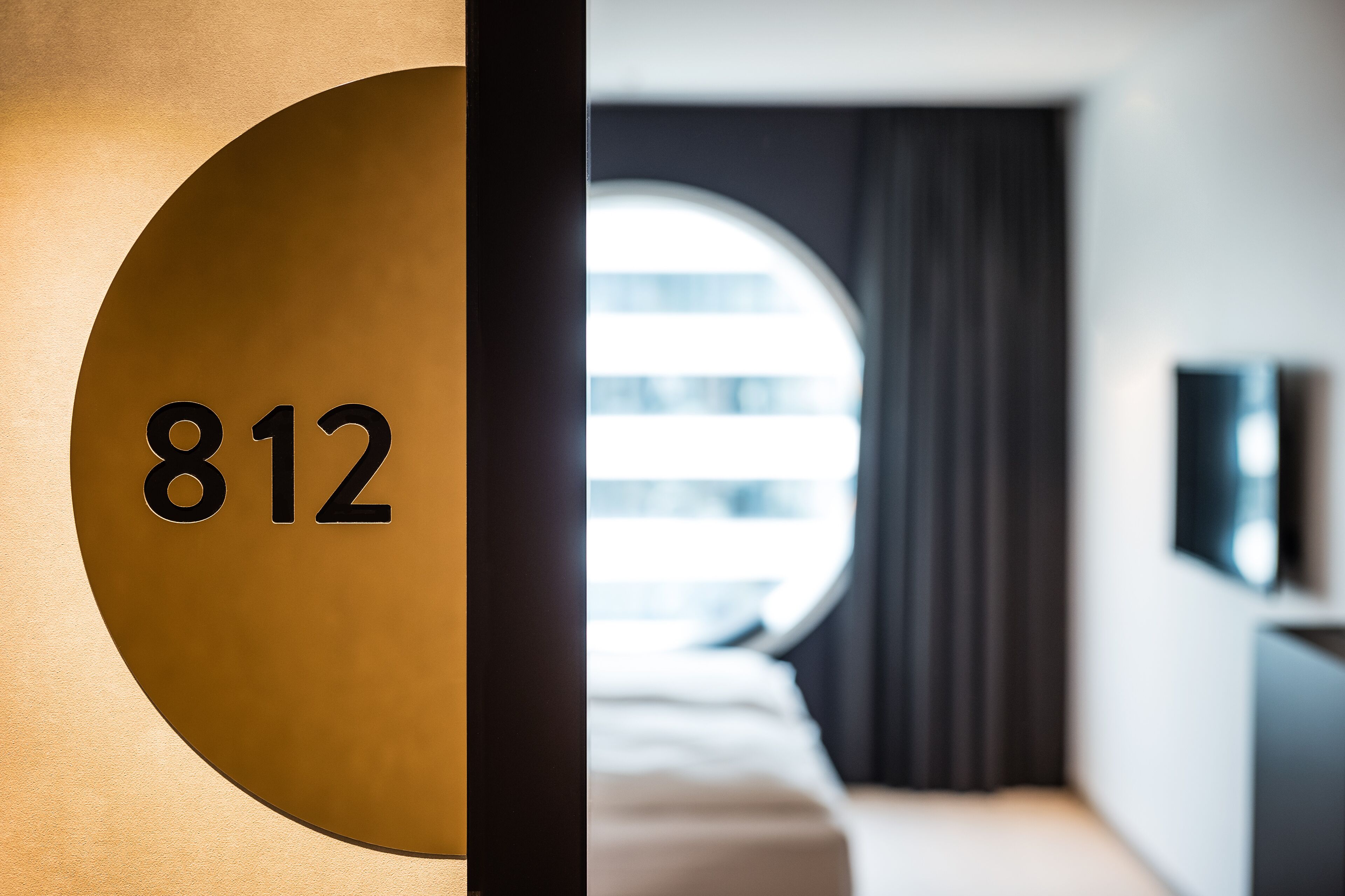 A hotel room with a large circular window, a bed, a TV mounted on the wall, and a door with the number 812 visible.
