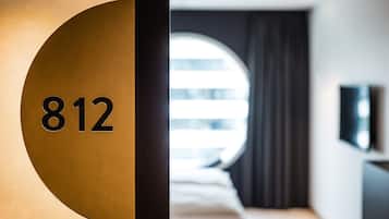 A hotel room with a large circular window, a bed, a TV mounted on the wall, and a door with the number 812 visible.