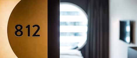 A hotel room with a large circular window, a bed, a TV mounted on the wall, and a door with the number 812 visible.