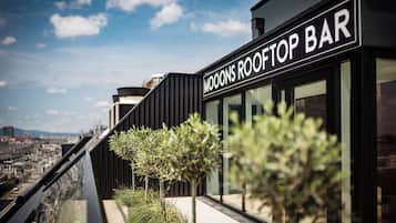 Un bar en la azotea llamado "MOOONS ROOFTOP BAR" con un diseño moderno, amplios ventanales y vistas panorámicas de la ciudad.
