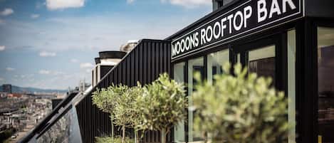Un bar sur le toit appelé « MOOONS ROOFTOP BAR », doté d’un design moderne, de grandes fenêtres et offrant une vue sur le paysage urbain.