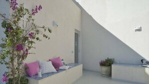 A sunlit outdoor seating area with a white wall, a potted plant, and a bench with cushions. - Community Rooms & Wine (Favara)
