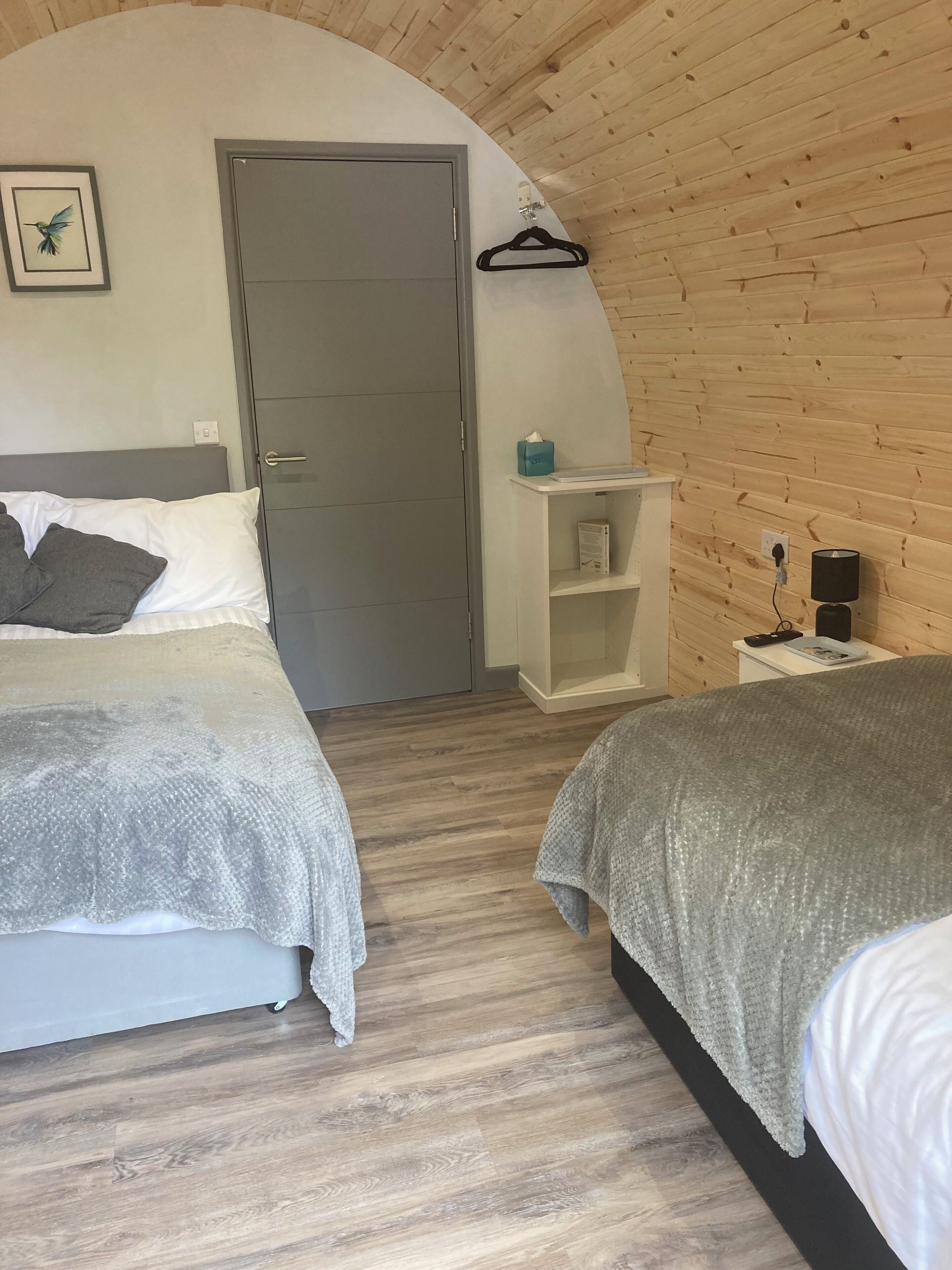 A bedroom with two beds, a wooden ceiling, a door, a small white shelf, and a wall-mounted lamp.