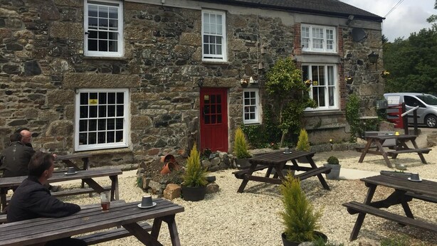 A stone building with a red door and multiple windows. There are outdoor wooden picnic tables and potted plants. - The Pirate Inn (Penzance)