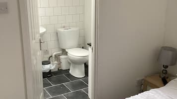 Une chambre d’hôtel avec un lit, une salle de bain aux carreaux blancs, des toilettes et une table de chevet avec une lampe.