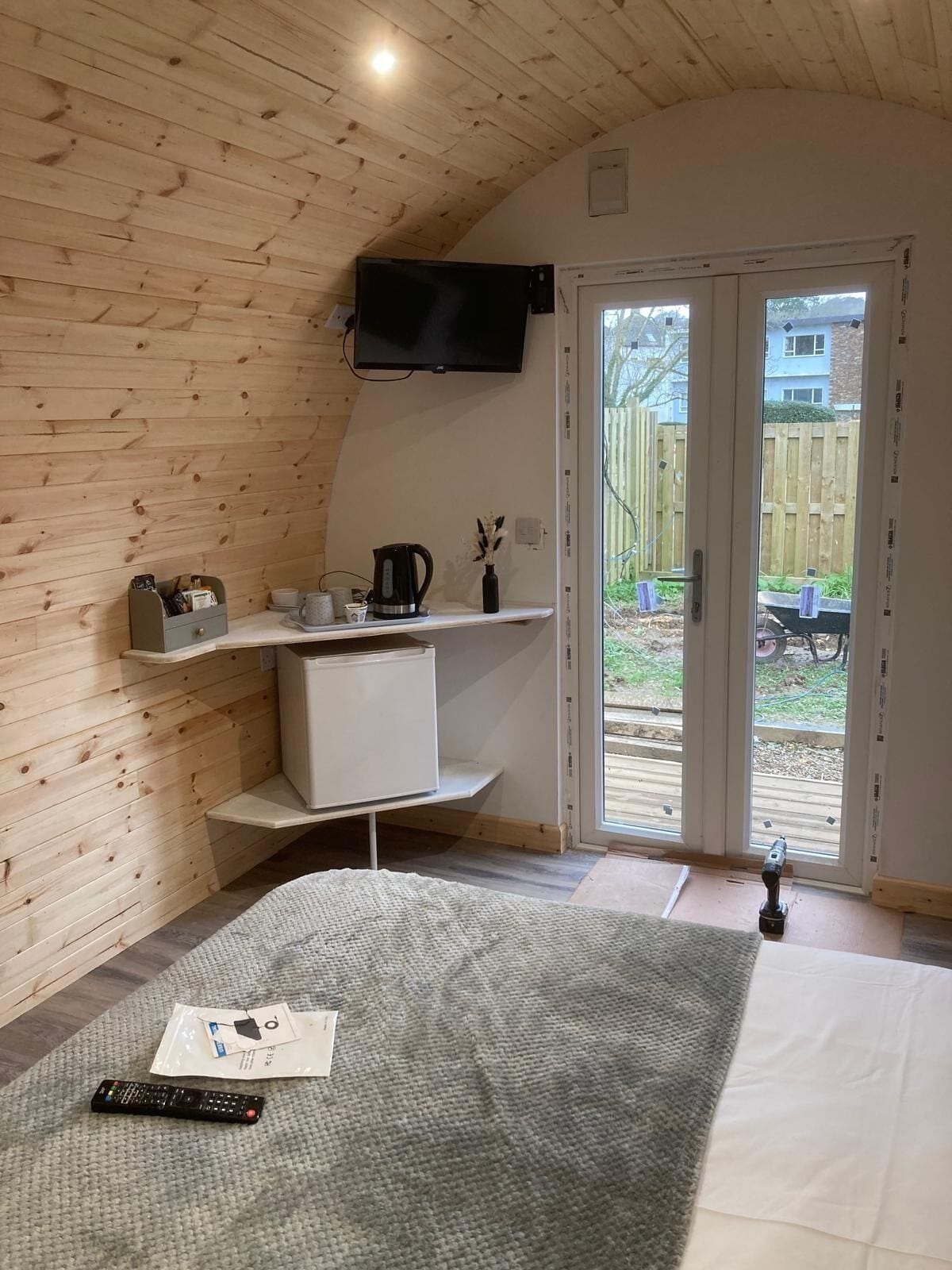 Ein Zimmer mit Holzbalkendecke, einem an der Wand befestigten Flachbildfernseher, einem weißen Schrank, einer Kaffeemaschine und einer Fernbedienung auf einem grauen Teppich.