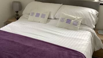 A neatly made bed with white linens and purple pillows, a bedside lamp, and a purple throw blanket.