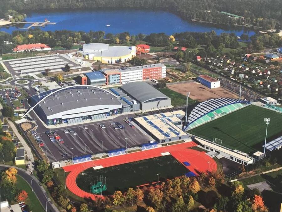 Vista aérea de un complejo deportivo con un estadio, zonas de estacionamiento y edificios adyacentes.