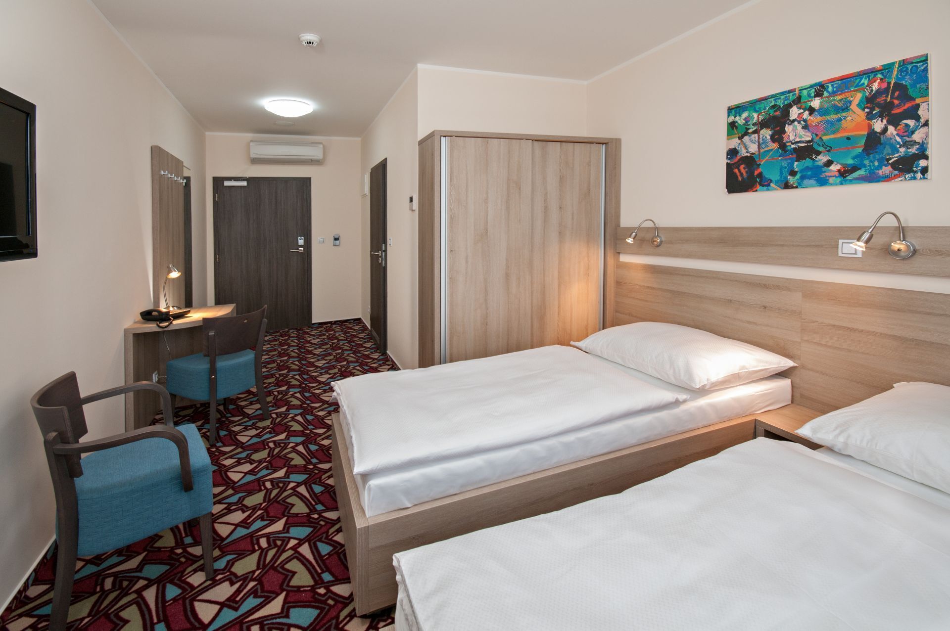 Hotellihuone, jossa on kaksi sänkyä, työpöytä tuolilla, suuri peili ja värikäs abstrakti taulu seinällä.