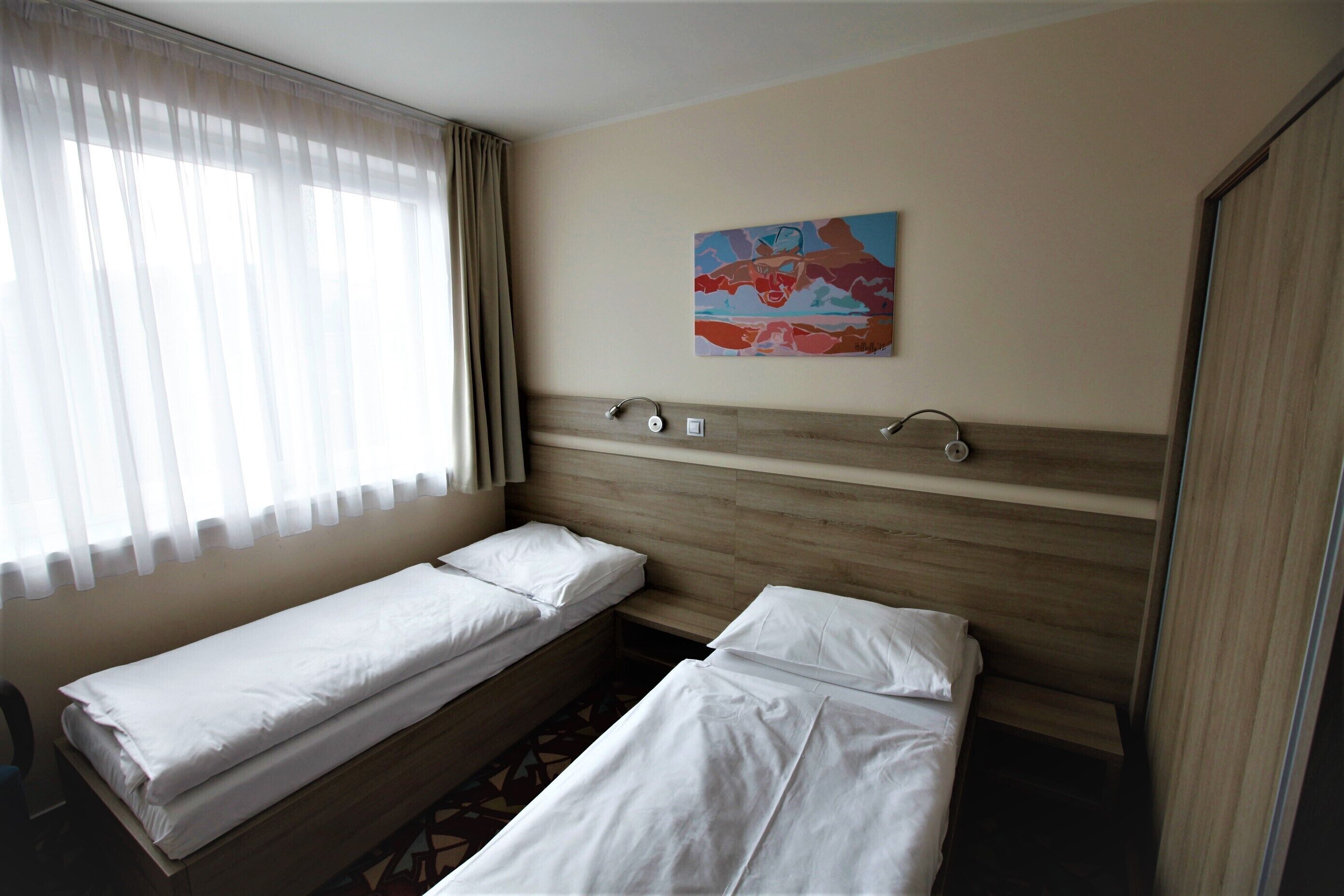 Habitación de hotel con dos camas individuales, cabecero de madera, ventana con cortinas traslúcidas y un cuadro abstracto de colores en la pared.