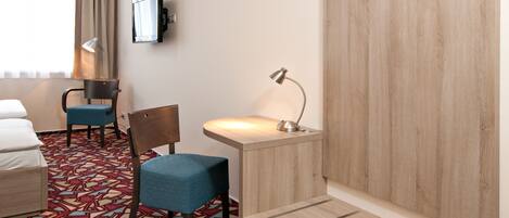 A hotel room with a bed, a desk, a chair, a desk lamp, a wall-mounted TV, and a patterned carpet.