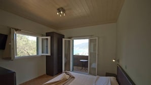 A bedroom with a bed, a TV, a balcony with a table and chairs, and a view of the outdoors. - Lefkas Petra (Lefkada)