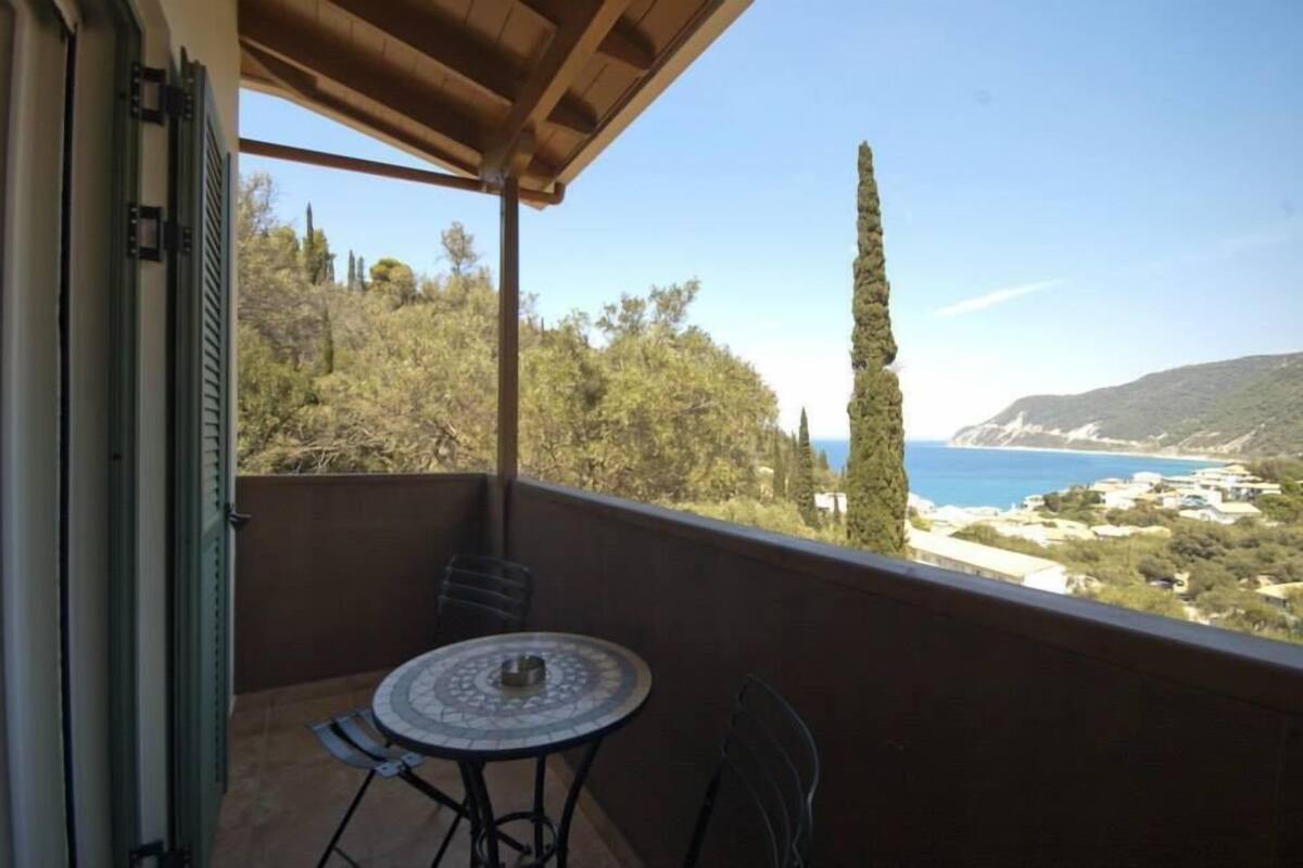 A balcony with a table and chairs, overlooking a coastal landscape with trees and a clear sky.