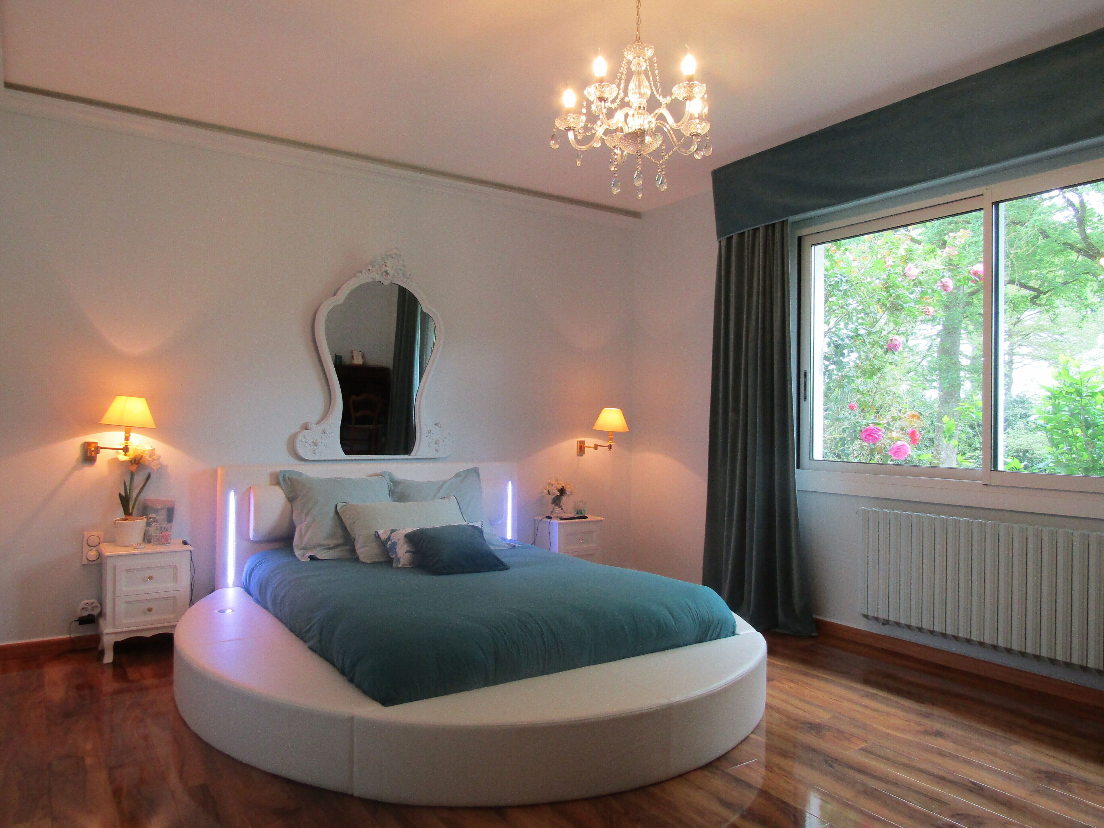 A bedroom with a round bed, two bedside tables, a mirror, and a window with a view of greenery.
