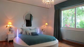 A bedroom with a round bed, two bedside tables, a mirror, and a window with a view of greenery.