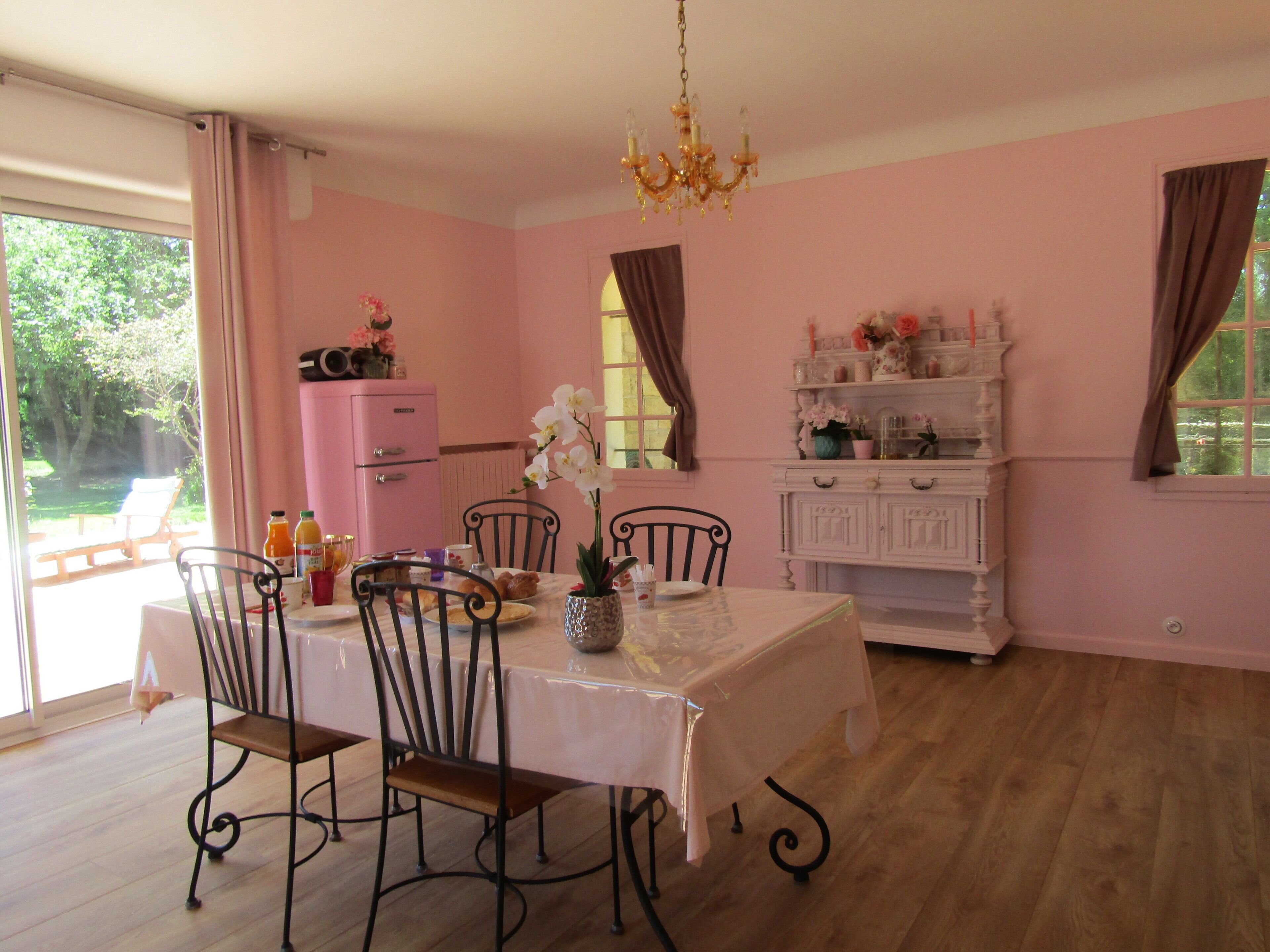 A dining room with a table set for four, surrounded by chairs. A pink refrigerator and a white cabinet with decorative items are visible in the background.