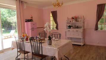 A dining room with a table set for four, surrounded by chairs. A pink refrigerator and a white cabinet with decorative items are visible in the background.