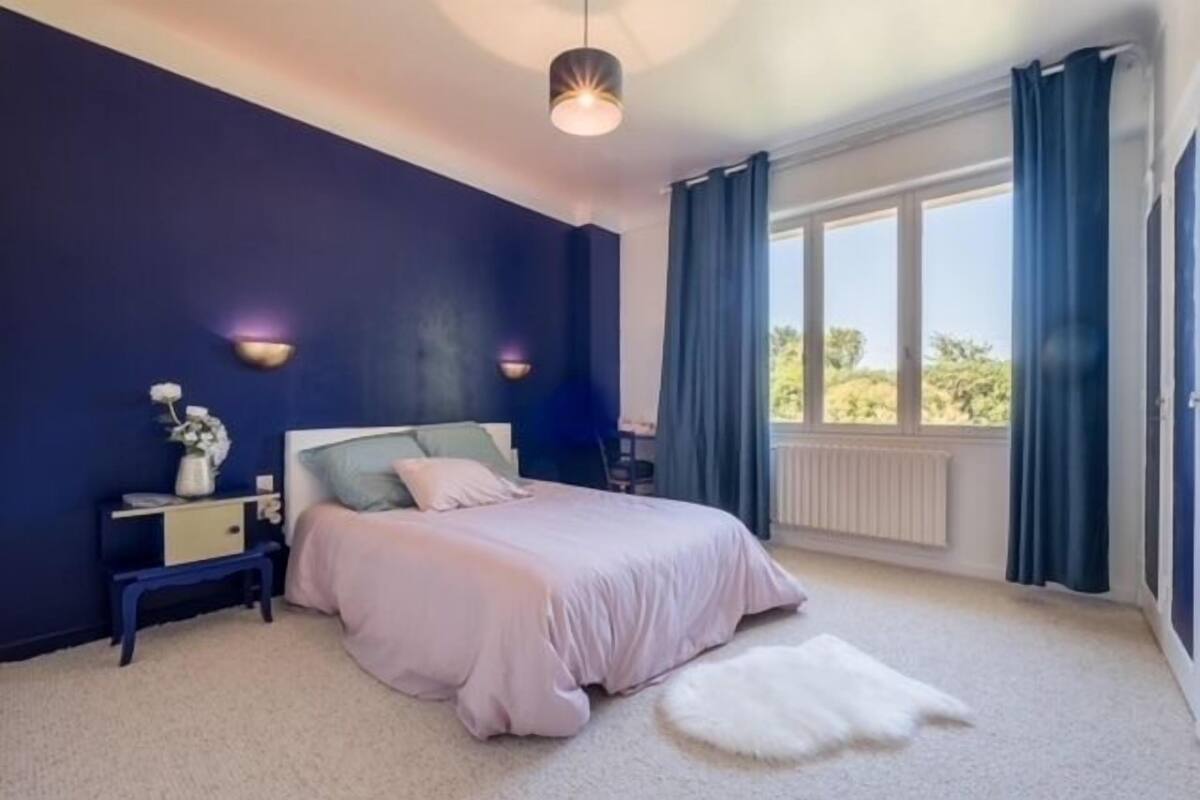 A bedroom with a bed, bedside tables, a nightstand, a window with curtains, and a view of greenery outside.
