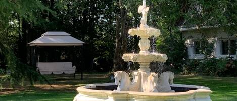 A multi-tiered stone fountain in a garden with a gazebo and a bench in the background.