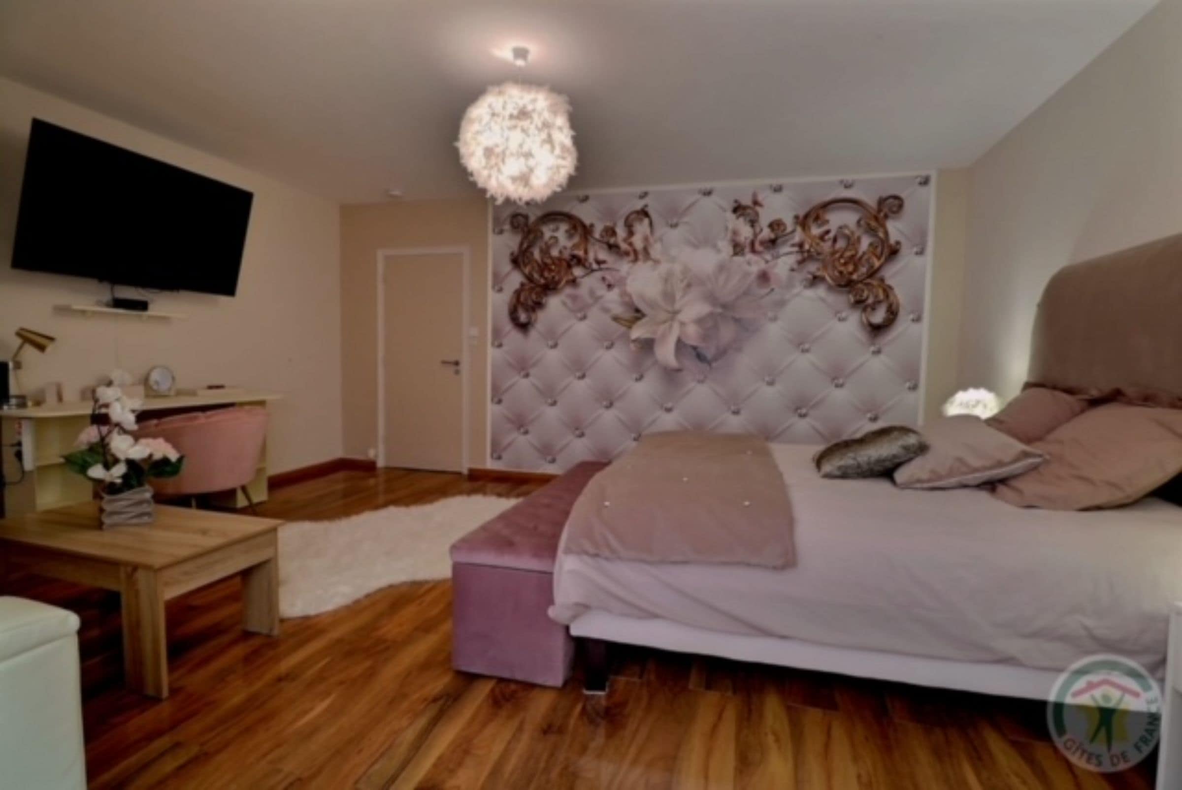 Un dormitorio con cabecera capitonada, una cama con almohadas, un televisor montado en la pared, piso de madera y una pequeña mesa con un jarrón de flores.