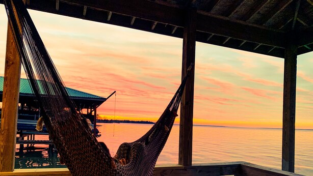 A hammock on a wooden deck overlooking a calm body of water during sunset.