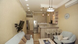 Una sala de estar moderna con un sofá, un sillón y una mesa de centro. Se ve una cocina con refrigerador y comedor al fondo.