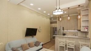 Una sala de estar con sofá, mesa de centro y televisor montado en la pared. Hay una zona de cocina con refrigerador y lavaplatos.