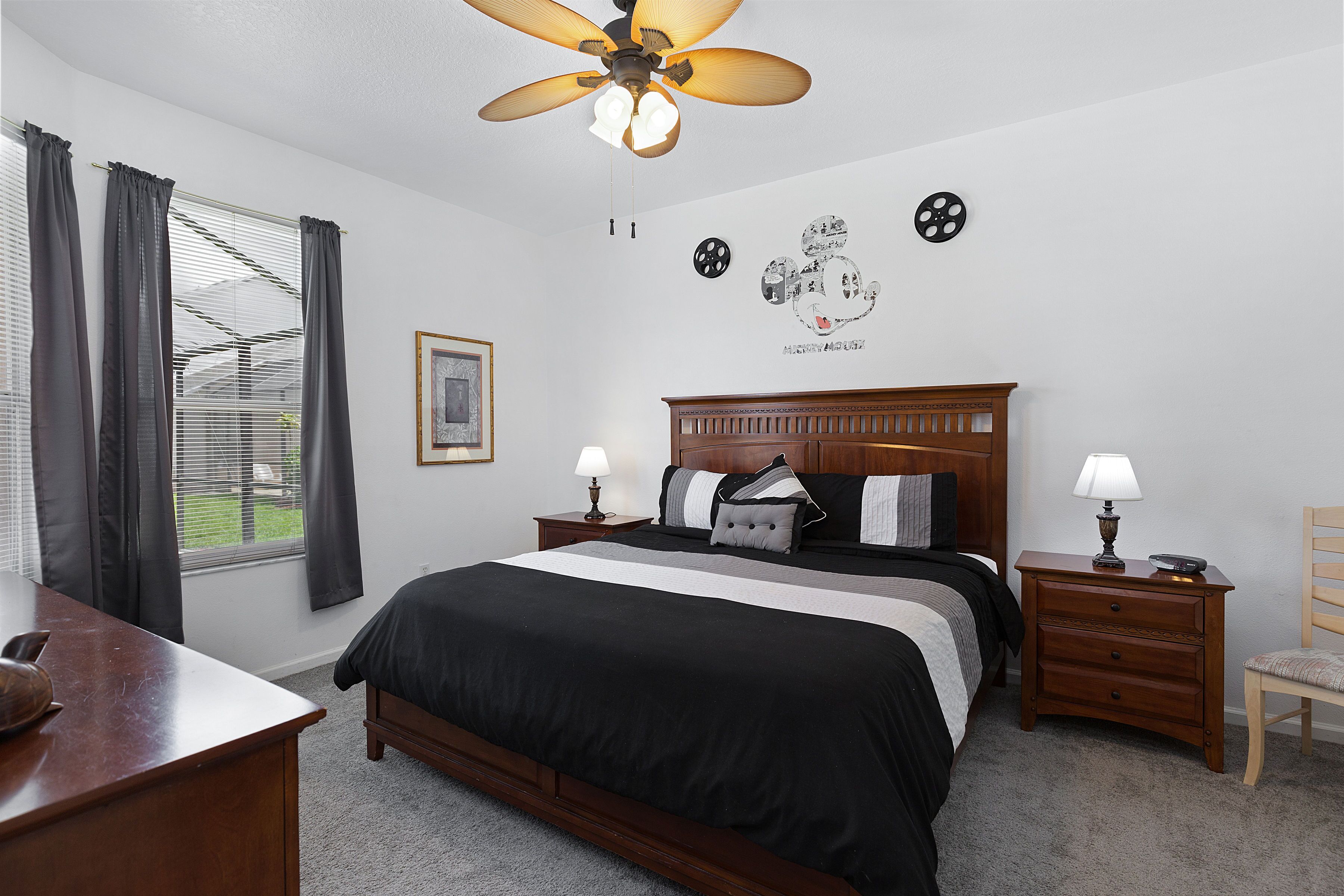 Une chambre à coucher comprenant un lit, des tables de chevet, un bureau, une chaise, un ventilateur de plafond et une fenêtre avec des rideaux.