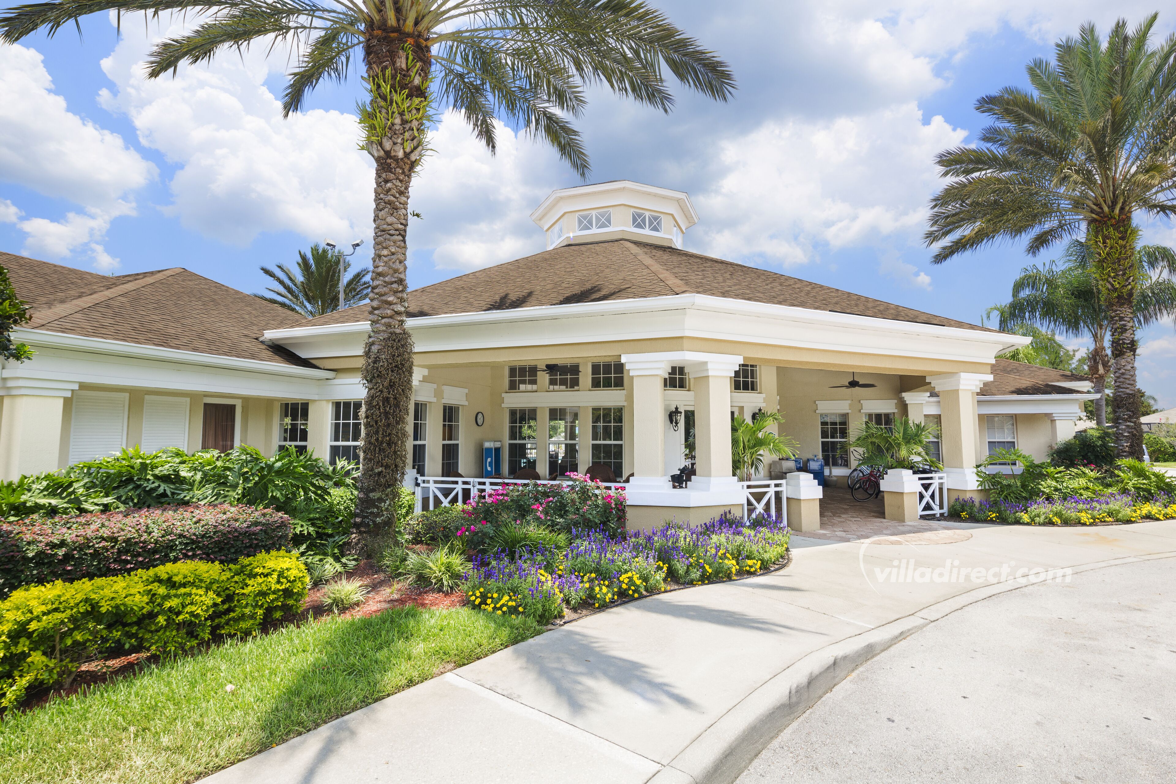 A two-story building with a covered porch, palm trees, and a variety of colorful flowers.