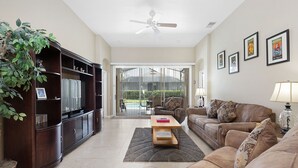A living room with a dark wooden entertainment unit, a brown leather sofa, a wooden coffee table, and a ceiling fan. - Our Happy Place by VillaDirect (Kissimmee)