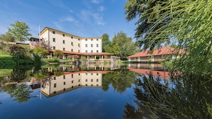 Ein Gebäude mit Spiegelung in ruhigem Wasser, umgeben von Grünflächen und Bäumen.