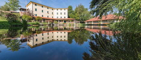 A building with a reflection in a calm water body, surrounded by greenery and trees.