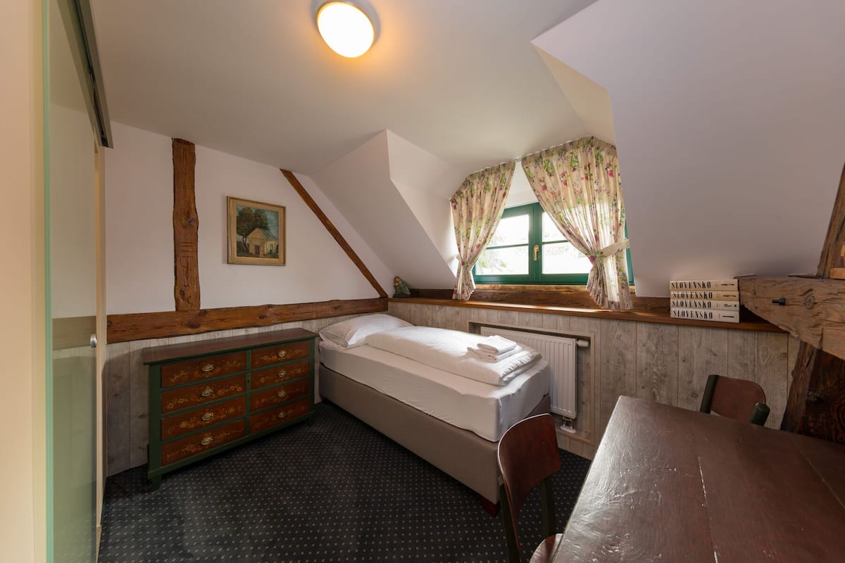 Una habitación pequeña y acogedora con una cama individual, un aparador de madera, una mesa y una ventana con cortinas.