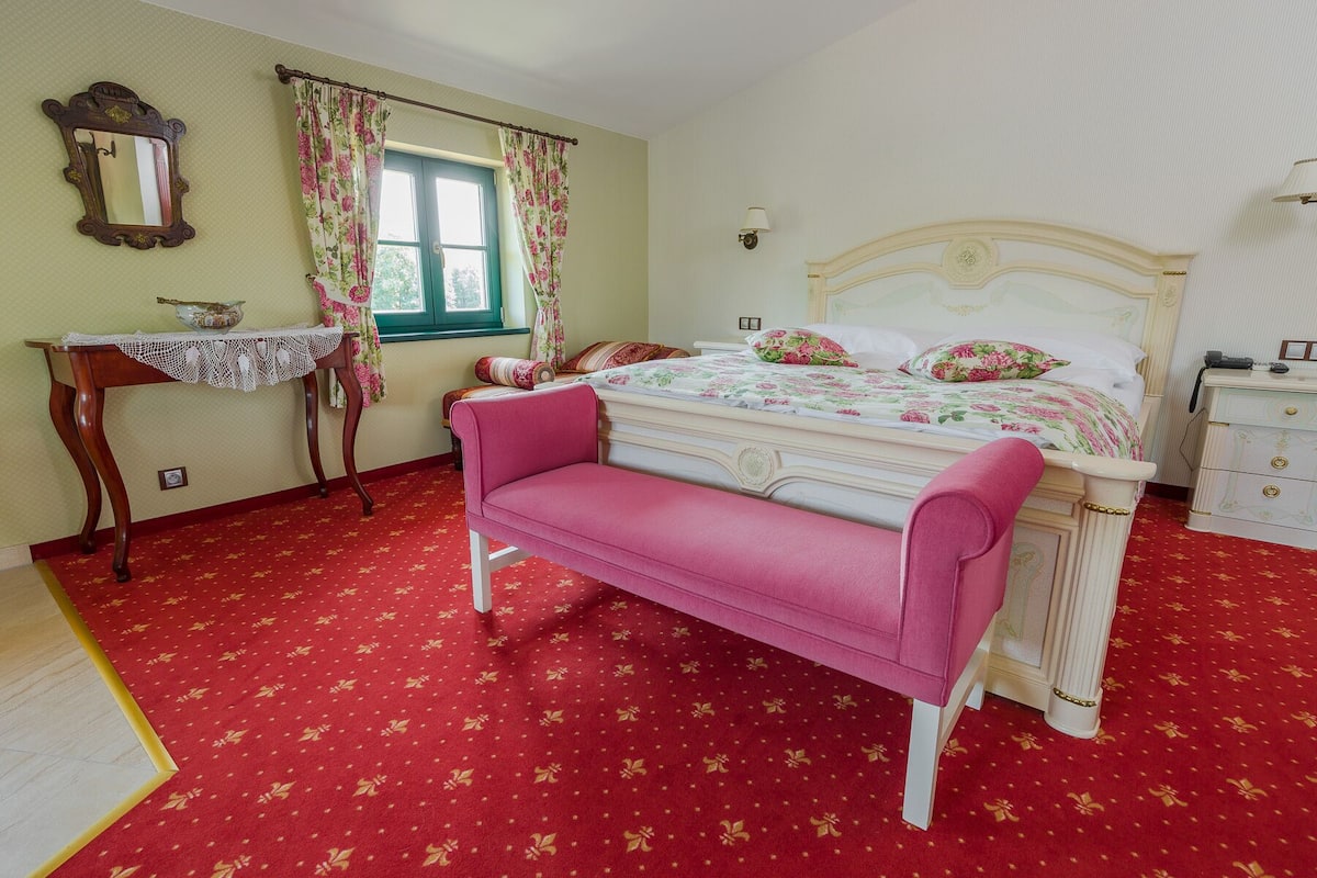 Un dormitorio con cama, banca, mesita de noche, lámpara y ventana con cortinas.