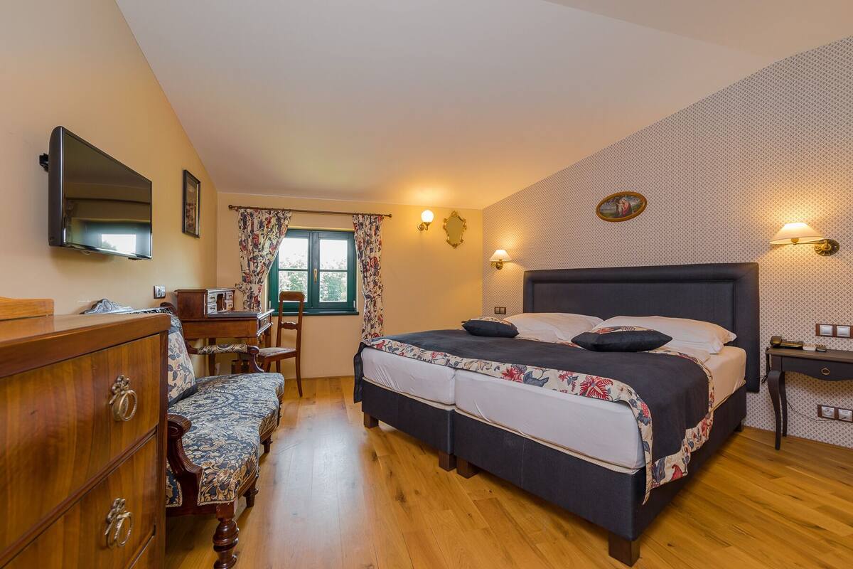Un dormitorio con una cama grande, un aparador de madera, un escritorio y una silla. Hay una ventana con cortinas, un televisor montado en la pared y dos apliques.